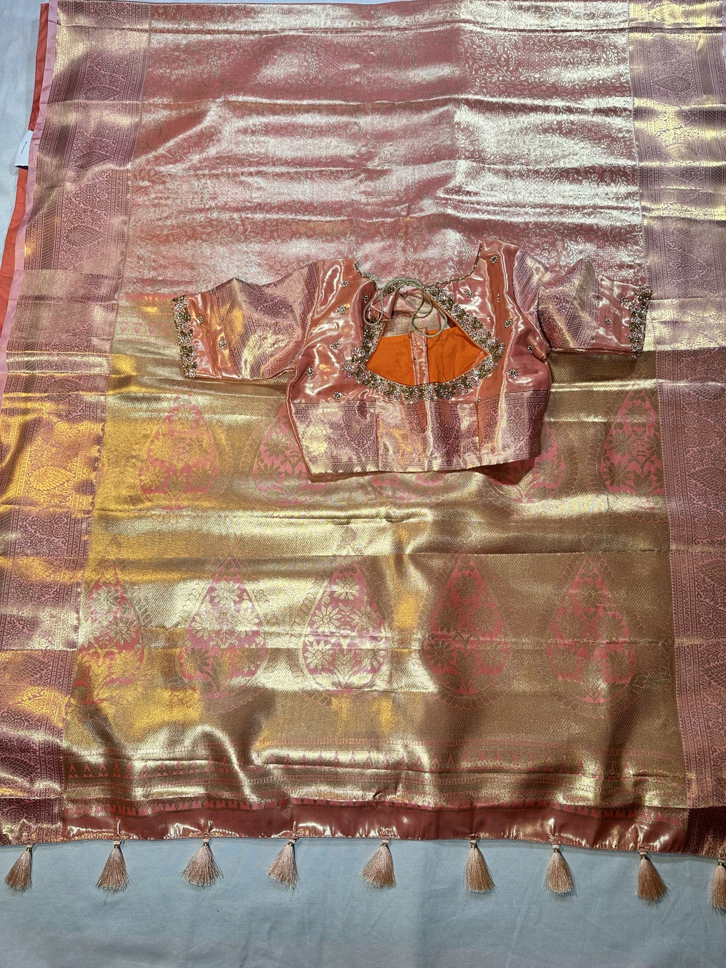 Kanjivaram semi silk saree with maggam work louse pastel shade
A pink and gold zari silk saree with a matching embroidered blouse piece featuring orange center panels, laid flat on a light blue-gray fabric background.