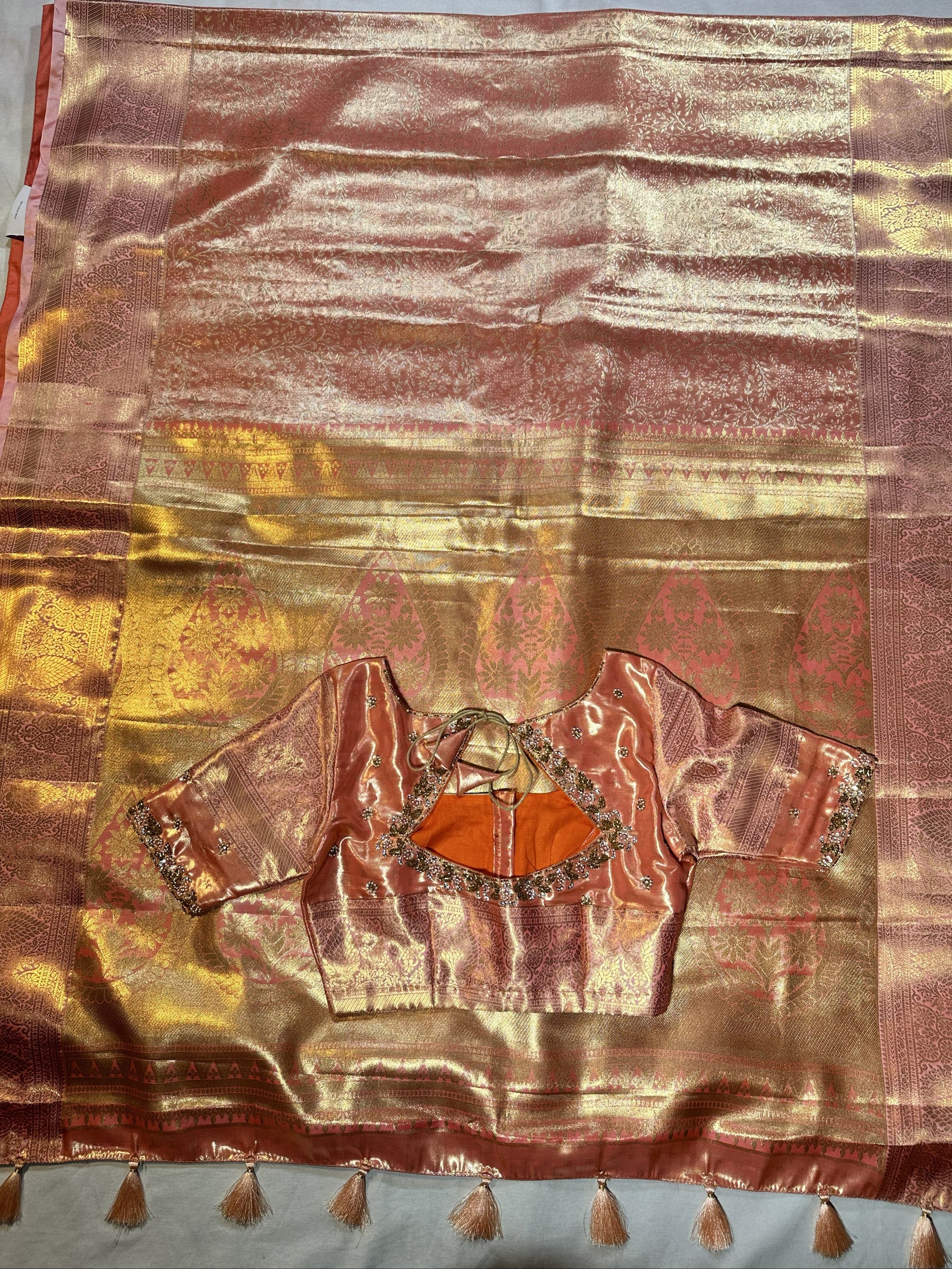 Kanjivaram semi silk saree with maggam work louse pastel shade
A pink and gold zari silk saree with a matching embroidered blouse piece featuring orange center panels, laid flat on a light blue-gray fabric background.