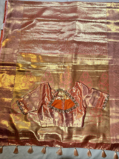 Kanjivaram semi silk saree with maggam work louse pastel shade
A pink and gold zari silk saree with a matching embroidered blouse piece featuring orange center panels, laid flat on a light blue-gray fabric background.